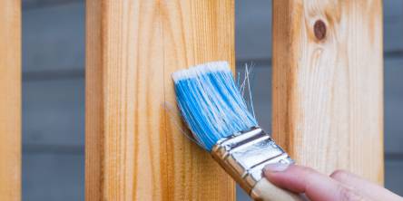 Man painting the slats of a wooden fence with a large paintbrush that has been dipped in blue paint.